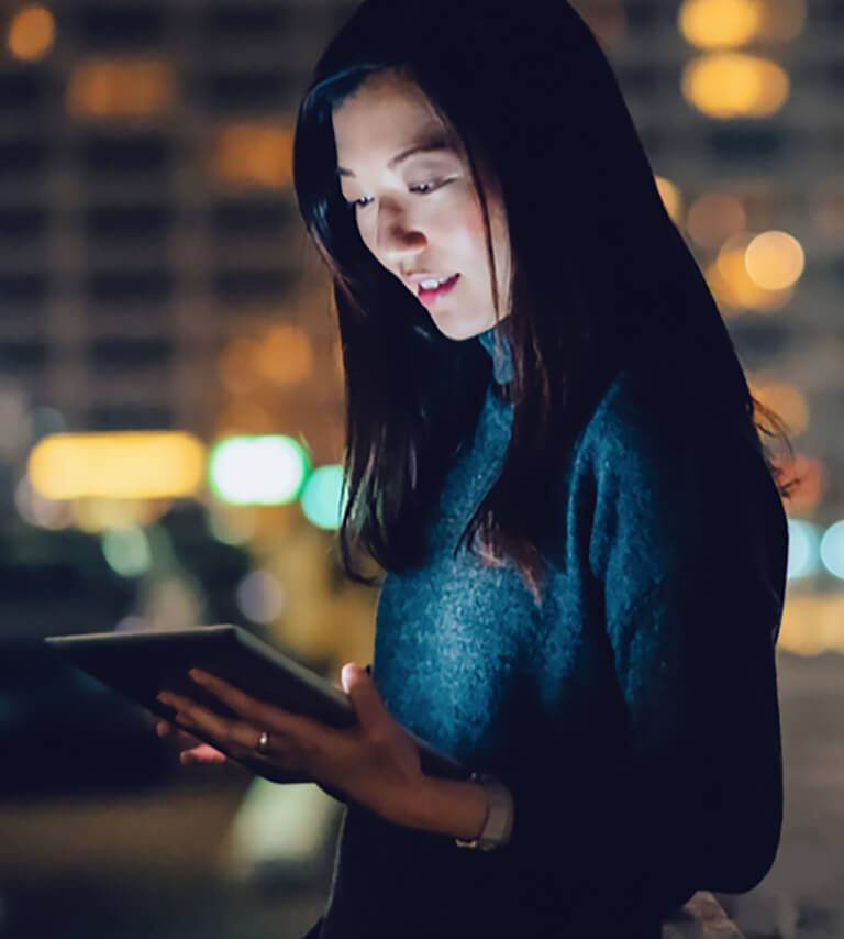 Als Beispiel für die Bildsprache ist das Foto einer jungen Dame zu sehen, die vor einer nächtlichen Stadtkulisse auf ein Tablet blickt